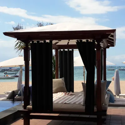 A cabana with black curtains and cushions on a beach with the ocean and sky in the background.