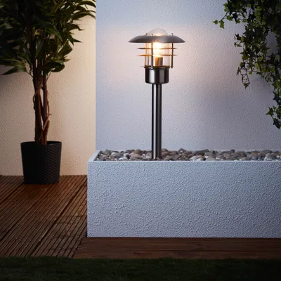 An outdoor lamp is shown in a planter box filled with stones, next to a potted plant and a wooden deck.