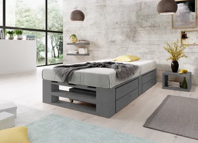 A bedroom with a modern, industrial-style bed frame made of grey wood, featuring built-in drawers and open shelving. The bed is dressed with grey bedding and a yellow pillow. The room has a textured grey wall, large windows with a view of trees, and minima