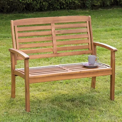 A wooden garden bench sits on a grassy lawn with a hedge in the background. A mug and saucer are placed on the bench.