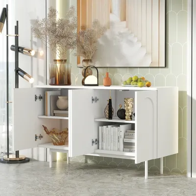A white sideboard with its doors open, displaying shelves with books and decorative items. The sideboard is placed in a room with a patterned wallpaper and a large artwork on the wall.