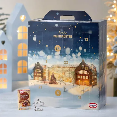 A Christmas advent calendar with a winter scene illustration, placed on a table with decorative paper houses and cookie cutters.
