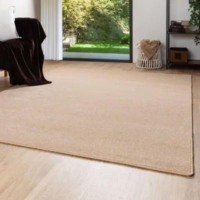 A living room with a large beige rug, a sofa with a brown blanket and white pillow, and a large window overlooking a garden.