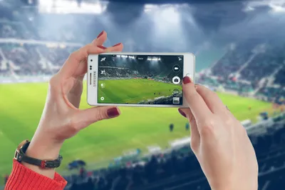 Two hands holding a smartphone, taking a picture of a soccer field in a stadium.