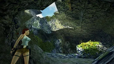 Lara Croft stands in a cave opening, looking out at a rocky landscape with vegetation.