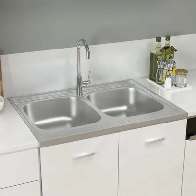 A stainless steel double sink with a faucet, set into a white countertop and cabinets, with some kitchen items in the background.