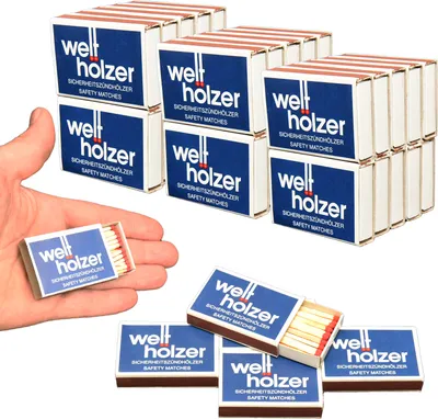 A hand holding a small box of matches with several other boxes stacked in the background.