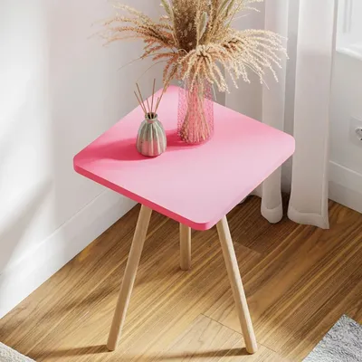 A pink side table with dried flowers and a diffuser on it, placed on a wooden floor next to a window with white curtains.