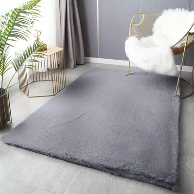 A plush gray rug is laid out on a light gray floor in a modern living room setting. A gold wire chair with a white fluffy throw is placed on the rug, and a small gold hexagonal side table with a plant and a decorative deer figurine is next to the rug.