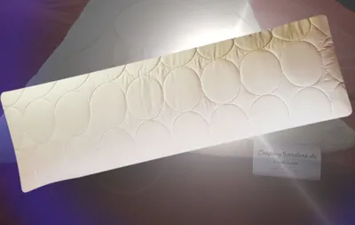 A cream-colored body pillow with a circular quilted pattern is laid out on a surface with purple and light gray lighting.