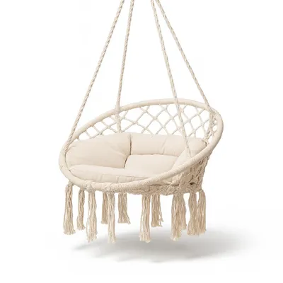 A beige macrame hanging chair with tassels and a cushion, suspended against a white background.
