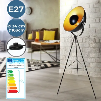 A black and gold tripod floor lamp is shown in a living room setting, with a sofa and coffee table in the background.