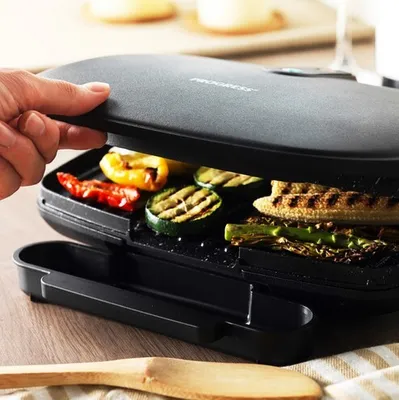 A hand is opening a black electric grill with vegetables inside, placed on a wooden table.