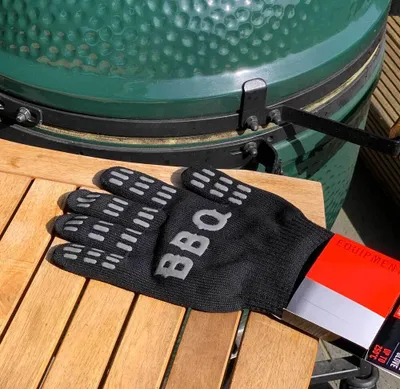A black BBQ glove with 'BBQ' written on it lies on a wooden table next to a green grill.