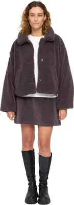 A woman is standing and modeling a brown faux-fur jacket and skirt set with black boots.