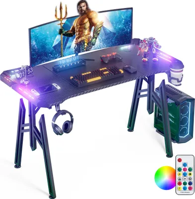 A gaming desk setup with a monitor, keyboard, mouse, controllers, PC tower, headphones, and RGB lighting.