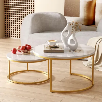 A modern living room setting with two round coffee tables, one nested inside the other, featuring marble-like tops and gold metal frames. The tables are adorned with decorative items like vases, a coffee cup, and fruit.