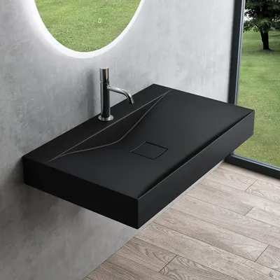 A modern black matte washbasin mounted on a wall, with a chrome faucet. The background shows a large circular mirror reflecting a green lawn and a window with a view of trees and grass.