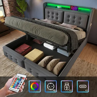 A grey upholstered bed with storage, featuring LED lighting and a remote control. The bed is shown with its storage compartment open, revealing items like luggage and pillows. Icons indicating features like USB charging and power outlets are displayed at t