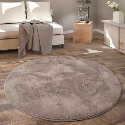 A round, shaggy rug is placed on a wooden floor in a living room setting, with a sofa and a side table visible in the background.