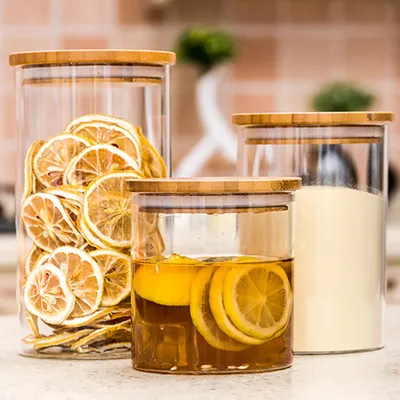 Three glass storage jars with bamboo lids are displayed on a countertop. The largest jar on the left is filled with dried lemon slices. The middle jar contains a liquid with lemon slices. The jar on the right is filled with a light-colored powder.