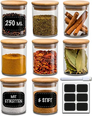 A collection of glass spice jars with wooden lids, arranged in a grid on a white background. Some jars have labels with text.