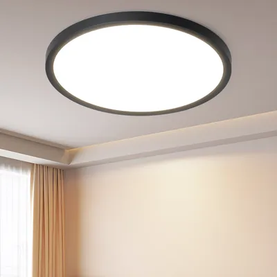 A modern, round LED ceiling light fixture with a black frame is mounted on a white ceiling in a room with beige walls and sheer curtains.