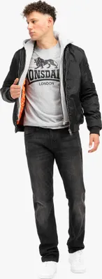 A man wearing a black bomber jacket over a gray Lonsdale t-shirt and dark wash jeans, standing against a white background.