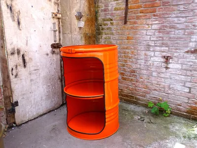 An orange barrel-shaped shelf unit is placed in an outdoor alleyway next to a weathered metal door and a brick wall.