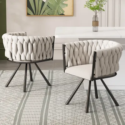 Two beige upholstered chairs with braided detailing and dark metal legs are placed on a patterned rug in front of a white dining table.