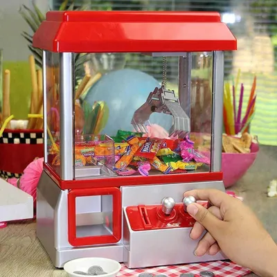 A hand is operating a candy claw machine filled with candy, with other party decorations in the background.