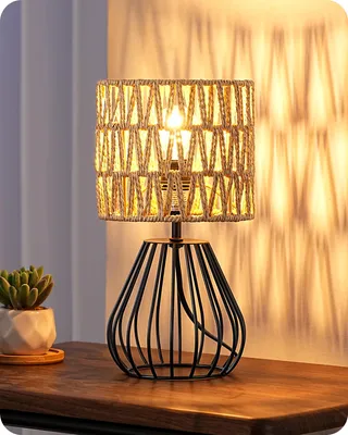 A table lamp with a woven shade sits on a wooden surface next to a small potted plant.