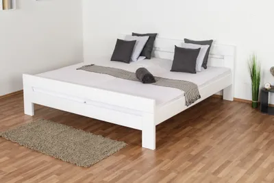 A white bed with pillows and a throw blanket is shown in a room with wooden floors and a small rug.