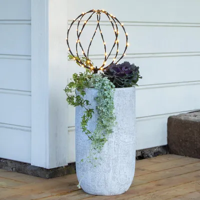 A decorative planter with plants and a lighted sphere on a wooden deck next to a white wall.