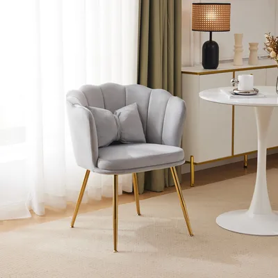 A light gray velvet chair with gold legs and a small decorative pillow sits in a room with sheer curtains and a white table.