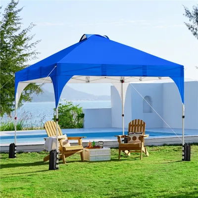 A blue pop-up canopy tent set up on a grassy lawn next to a swimming pool, with two wooden chairs and a small table with drinks.