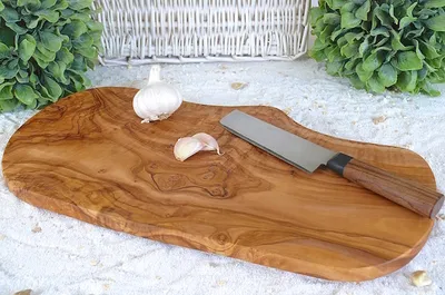 A rustic olive wood cutting board with a knife, garlic bulb, and a garlic clove on a white textured surface, with greenery and a basket in the background.