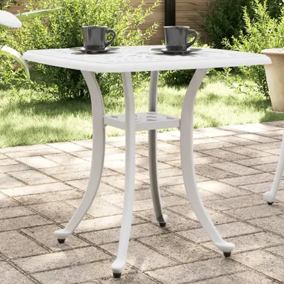 A white square garden table with two cups of coffee on it, set on a patio with greenery in the background.