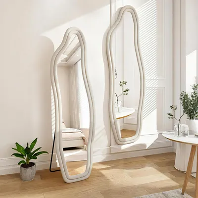 Two full-length mirrors with wavy frames are placed in a well-lit room with sunlight streaming in. One mirror reflects a bedroom scene, while the other reflects a table with plants. A potted plant sits in the foreground.