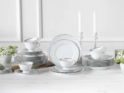 A table setting with a complete dinnerware set, including plates, bowls, and cups, arranged neatly. Two white candles in candle holders are in the background.