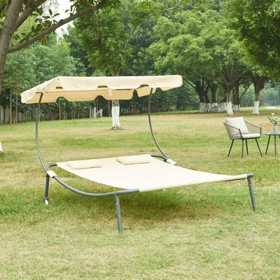 A double lounge chair with a canopy sits on a grassy lawn in a park-like setting with trees in the background.