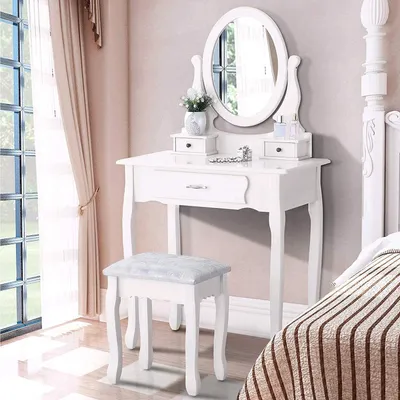 A white vanity table with an oval mirror and a matching stool is set up in a bedroom next to a window with sheer curtains and a bed with striped bedding.