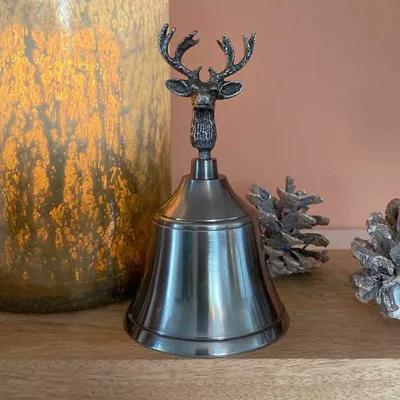 A decorative bell with a reindeer head on top, placed on a wooden surface next to a large candle holder and pinecones.