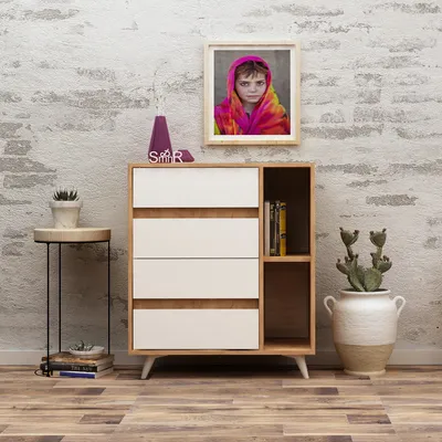 A modern dresser with white drawers and wooden accents, placed against a textured white brick wall. A framed picture of a child wearing a colorful headscarf hangs above it. Decorative items like a vase, books, and a potted cactus are placed around the dres