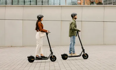 Two people riding electric scooters on a paved walkway in front of a modern building.
