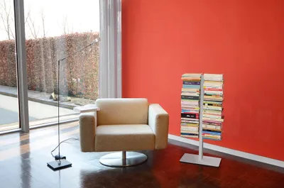 A modern living room with a beige armchair, a floor lamp, and a silver bookshelf filled with books, set against a vibrant red wall.