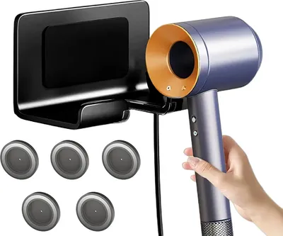 A hand holding a blue and gold hair dryer next to a black wall-mounted holder.