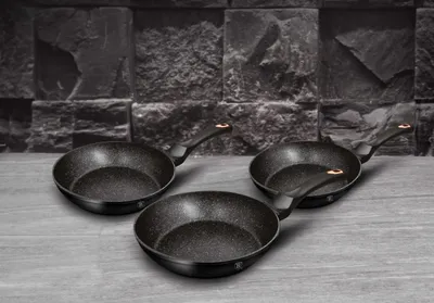 Three black frying pans with speckled interiors and gold accents on the handles are arranged on a gray surface in front of a dark, textured stone wall.
