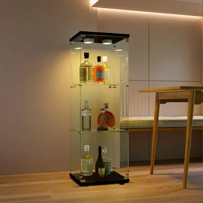 A glass display cabinet with bottles of alcohol on shelves, placed in a room with a table and bench.