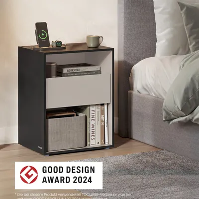 A modern black and gray nightstand with a charging phone, a mug, books, and a storage basket, placed next to a bed.